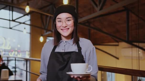 フレンドリーなアジアの女性ウェイトレスがコーヒーカップを持ってカメラを見ています 陽気で美しいバリスタ カフェスタッフのコンセプト — ストック動画