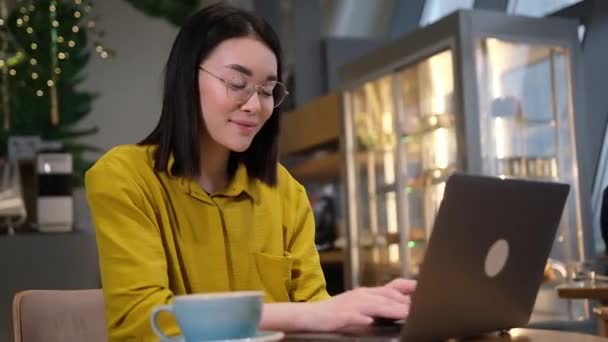 カフェでラップトップを使用するアジアの女性 コーヒーショップに座ってコンピュータで作業している若い美しい女の子 — ストック動画