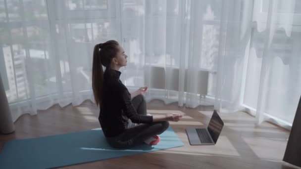 若い穏やかな女性がスタッフに座っている自宅のインテリアでラップトップの近くに簡単にハスのポーズ 目を閉じて床の上に一人で瞑想 オンラインヨガのトレーニング 午前中に呼吸運動を行う ストレスなし — ストック動画
