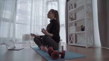 Genç sakin bir kadın evde dizüstü bilgisayarın yanında rahat bir şekilde oturuyor, yerde tek başına meditasyon yapıyor gözleri kapalı, internetten yoga eğitimi alıyor, sabahları nefes alma egzersizi yapıyor, stres yok.