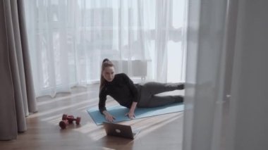 Evde dizüstü bilgisayarla antrenman yapan genç ve güzel bir kadın. Ayaklarını paspasın üstüne koy. Yoga, pilates, egzersiz.