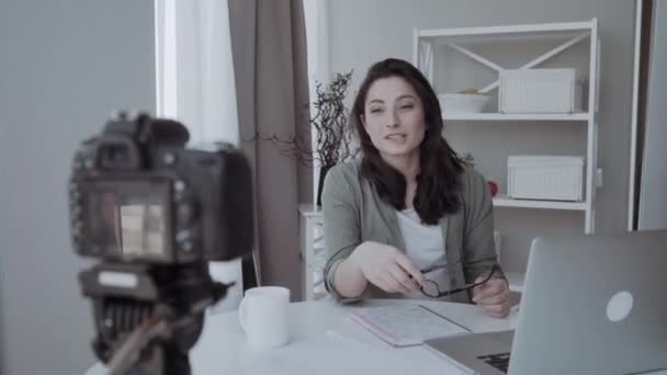 柔軟な三脚に取り付けられたデジタルカメラでビデオを録画する若い女性 笑顔の女性が机に座ってラップトップコンピュータで作業 — ストック動画