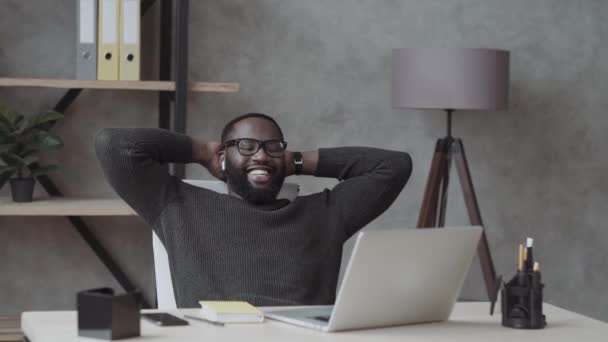 松懈的非洲商人头戴耳机喜欢听音乐手背坐在办公桌前 年轻员工休息一下感觉心情舒畅缓解压力的概念 — 图库视频影像