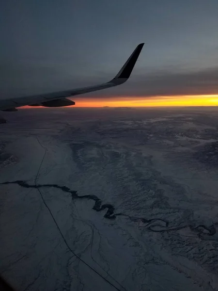 日落时透过飞机的窗户观看 云彩覆盖了整个天空 只有地平线上一片明亮的橙色 就在翼梢下方 下面的土地是山区 有一条蜿蜒的大河 — 图库照片