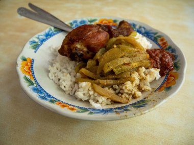 Bir parça kızarmış tavuk budu ve biber soslu pilav ve desenli beyaz tabak ve çatal bıçakla desenli krema renkli bir masada servis edilir..