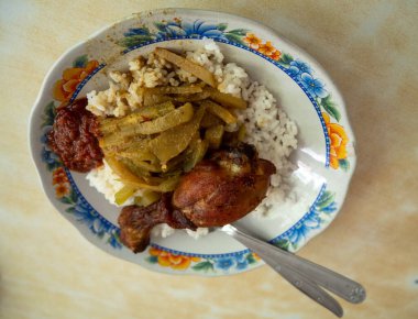 Bir parça kızarmış tavuk budu ve biber soslu pilav ve desenli beyaz tabak ve çatal bıçakla desenli krema renkli bir masada servis edilir..