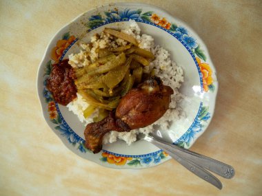 Bir parça kızarmış tavuk budu ve biber soslu pilav ve desenli beyaz tabak ve çatal bıçakla desenli krema renkli bir masada servis edilir..
