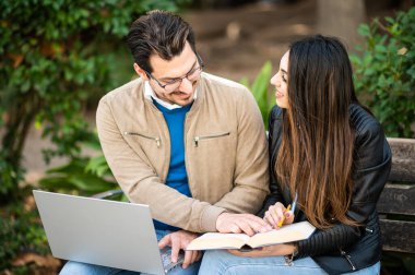 İki gülümseyen öğrenci açık bir bankta kitap ve dizüstü bilgisayar kullanarak oturuyorlar.