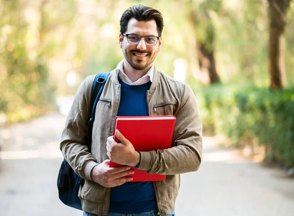 Erkek öğrenci bir üniversite parkında kameraya bakarak gülümsüyor.