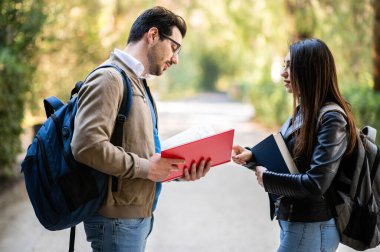 İki öğrenci, erkek ve kadın, ellerinde kitaplarla dışarıda konuşuyorlar.