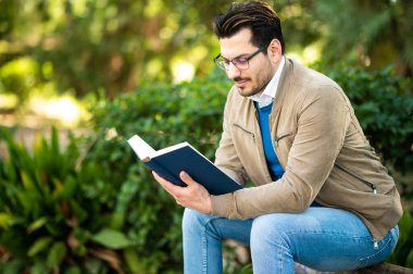 Erkek öğrenci kitap okuyor. Açık bir bankta oturuyor.