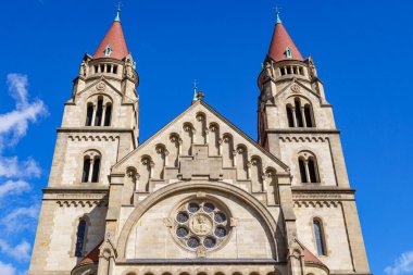 St. Francis Assisi Kilisesi cephesinin yakından görüntüsü, ikiz kuleler, kemerli pencereler, ve parlak mavi gökyüzünün altında dini imgeler içeren detaylı bir gül penceresi..