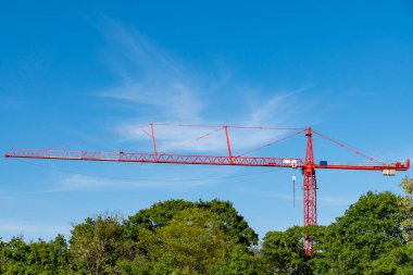 Uzun kırmızı bir vinç, açık mavi bir gökyüzüne karşı dimdik duruyor. Önünde yeşil ağaçlar var. Doğanın ve modern gelişimin ortasındaki inşaatı simgeliyor..