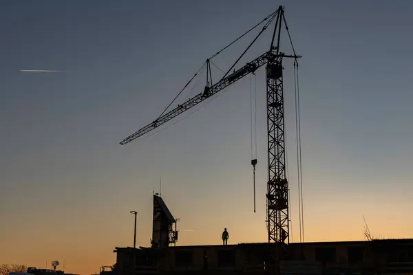 Gün batımında bir inşaat alanında çalışan vinç ve işçinin silüeti endüstriyel gelişimi ve dramatik bir akşam gökyüzüne karşı kentsel gelişimi gösteriyor..