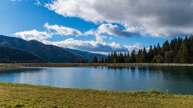 Sonbaharda Avusturya Alplerinde, parçalı bulutlu bir gökyüzünün altında, yeşil ağaçlar ve dağ yamaçlarıyla çevrili sakin alp gölü..