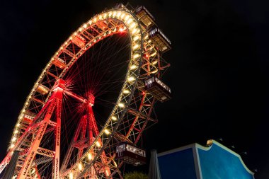 Geceleyin, bir lunaparktaki karanlık gökyüzüne karşı parlak kırmızı ve sarı ışıklarla aydınlanan dev dönme dolap..