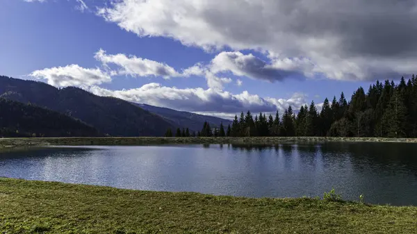 Sonbaharda Avusturya Alplerinde, parçalı bulutlu bir gökyüzünün altında, yeşil ağaçlar ve dağ yamaçlarıyla çevrili sakin alp gölü..