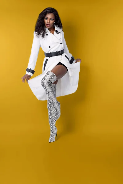 Full length of black female model in stylish white coat and thigh high snake skin high heel boots isolated posing on gold background