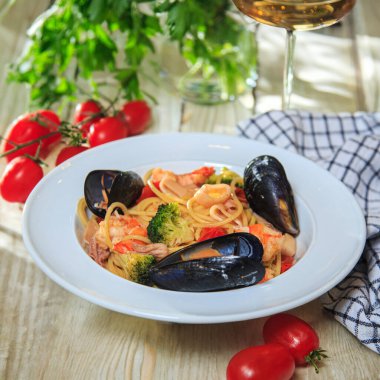 Spagetti makarnası, deniz ürünleri ve kiraz domatesli. Peçetedeki ahşap bir masada bir bardak şarapla servis ediliyor.