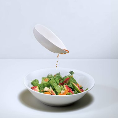 Siyah soslu beyaz bir sos teknesi beyaz bir kasenin üzerinde yeşil salata, incir ve peynir ile beyaz bir masada yüzüyor.