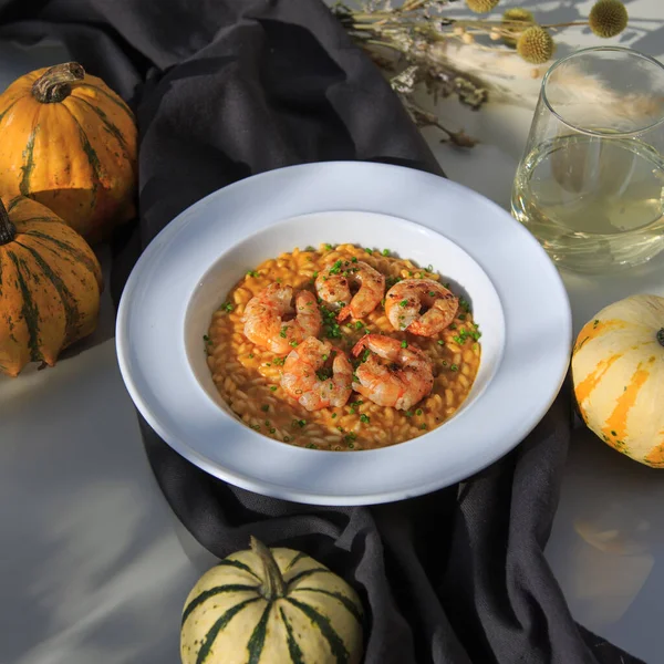 pumpkin risotto with grilled shrimp on a white plate on a gray tablecloth on a marble table with a glass of wine. pumpkins lie nearby in the sun's rays