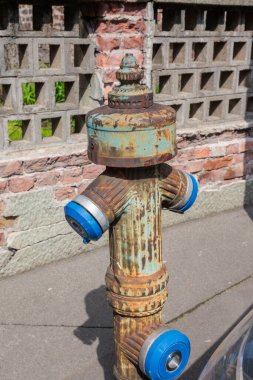 Görüntü caddeye kurulmuş bir yangın musluğunu gösteriyor. Yangın musluğu kırmızıya boyanmış ve yangın hortumlarını bağlamak için iki hortumu var. Yangın musluğunun kapağı kapalı ve vücut su kaynaklarını kontrol etmek için vanaları gösteriyor.. 