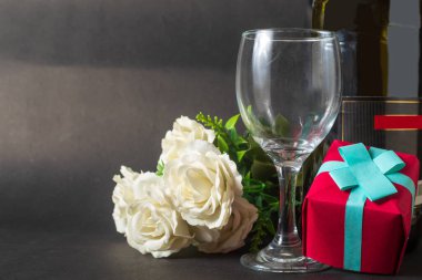 Sevgililer Günü kutlama geçmişi. Beyaz gül buketi, kırmızı hediye kutusu, kargo şişesi ve siyah arka planda şarap kadehi..