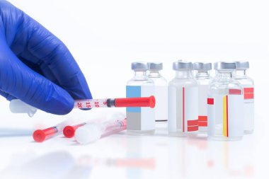 Mavi eldivenli doktor elinde şırınga ve şırınga destesiyle beyaz arka planda küçük şişelerle yatıyor.