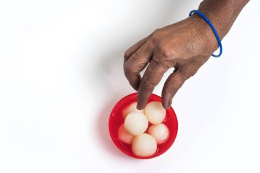 A hand with blue bracelet picks a traditional Indian sweet from a red bowl. clipart