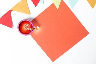 Bright Diwali oil lamp on top of orange paper with festive paper decorations on white background clipart