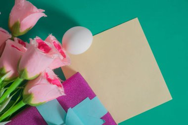 Renkli yeşil arka planda pembe güller, paketlenmiş hediye, yumurta ve boş kart içeren düz bir kompozisyon.