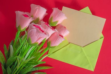 Kırmızı arkaplanlı yeşil bir zarfın yanına yerleştirilmiş pembe güller sevgiyi, takdiri ya da kutlamaları çağrıştırıyor..