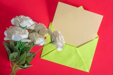 Parlak kırmızı bir yüzey üzerinde canlı yeşil bir zarfın yanında narin beyaz güllerin güzel bir düzeni..