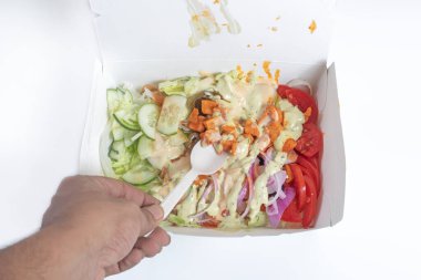 Salatalık, marul, domates, soğan ve kıtır ekmekten oluşan canlı bir salata, üzerine krema sosu konmuş, elinde plastik çatalla servis edilen bir paket servis konteynerinde servis ediliyor..
