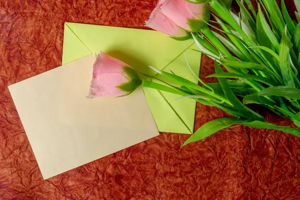 Yeşil bir zarfın içindeki pembe bir gül, boş bir kartla desenli bir kumaşa yerleştirilir, selamlama ya da iletişim teması için mükemmeldir..