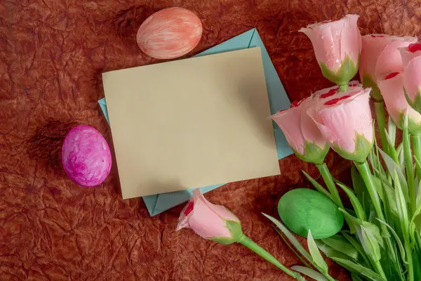 Güzel bir pembe gül aranjmanı, renkli Paskalya yumurtaları ve kırsal bir zemine kurulmuş boş bir kart. Bahar kutlamaları için mevsimlik temalar için mükemmel.