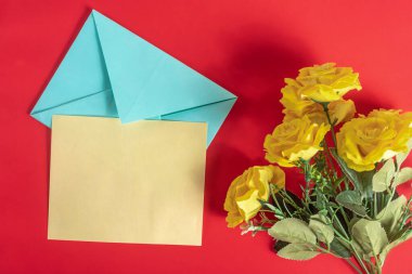 Açık bir zarf ve boş bir kağıdın yanına yerleştirilmiş canlı sarı güller, koyu kırmızı bir zemin üzerine kurulmuş, kutlama temalarını iletiyor, ve aşk.