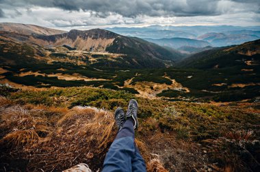 Dağların eteklerinde, yeşil doğa ve fırtınalı karanlık gökyüzü arasında oturan erkek bacakları kapalı. Seyahat ve turizm kavramı.