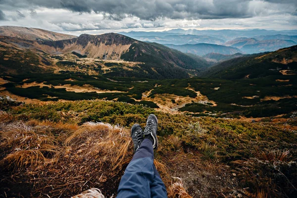 stock image Close up of male legs in hikking shoes sitting on the edge of mountains among green nature and stormy dark sky. Concept of travel and tourism.