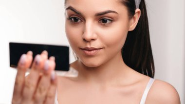 Güzel genç bayan aynaya bakıyor ve kozmetik güzellik tedavisinden zevk alıyor. Stüdyoda beyaz arka planda izole edilmiş.