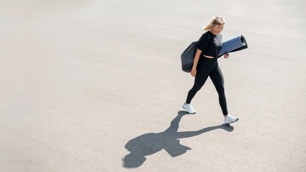 屋外でのトレーニングの準備をしながら 若い女性が通りを横断し ヨガマットを運ぶ笑顔 健康的なライフスタイルのコンセプト — ストック写真