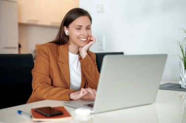 Gülümseyen, kulaklık takan genç bir iş kadını masasında, parlak, modern bir ofiste dizüstü bilgisayarla çalışıyor. Stok fotoğrafı