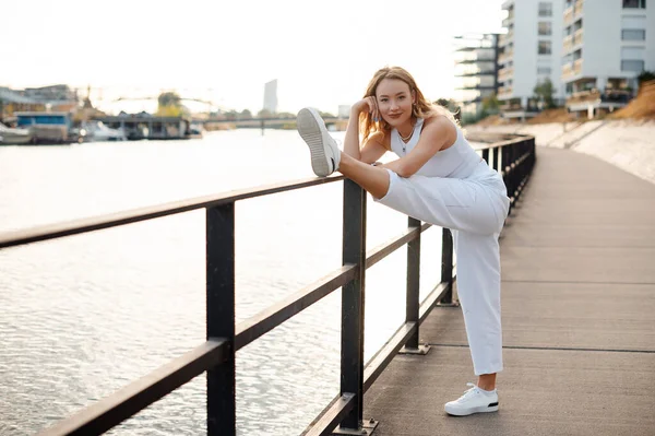 笑顔の若い女性の足を伸ばし 川の近くのカメラを見ての完全な長さのビュー 朝の屋外で運動フィットネスモデル — ストック写真