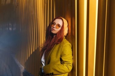 Şehirli duvar arkasında poz veren hippi kızıl saçlı kadın. Sokakta duran, güneş gözlüklü, kentsel giysili kızıl kız.