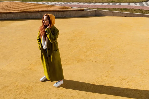 stock image Full length view of the ginger woman wearing stylish coat posing at the urban stadium. Red haired girl in sunglasses standing at the street