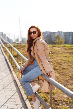 Kızıl saçlı, dişlek tebessümlü, sokakta oturan ve dışarıda yürüyen genç kızın dikey görüntüsü. Mutlu kadın kameraya bakıyor.