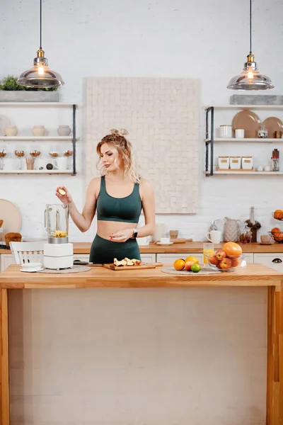 キッチンに立つ緑のスポーツ衣装でブロンドフィットネス女性の垂直ビューと新鮮なオレンジジュースをやっています 健康的な食生活の概念 — ストック写真