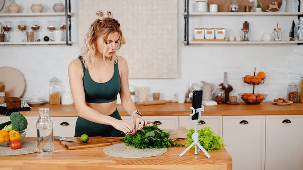 グリーンスポーツ衣装カットサラダで魅力的な強い女性と健康食品についてのモバイル上のビデオを記録します フィットネスブロガーとともにブロンド髪スタンディングオンキッチンと作るチュートリアル — ストック写真