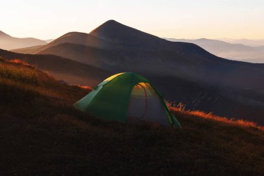 Dağın tepesinde yeşil kamp çadırı ve arka planda muhteşem bir gündoğumu. Temiz hava için zaman harcamak. Seyahat konsepti.