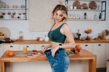 Kısa sarı saçlı, güzel ve sportif bir kadın. Parlak mutfakta poz veriyor. Büyük beden kotla. Kilo kaybı ve diyet konsepti. 